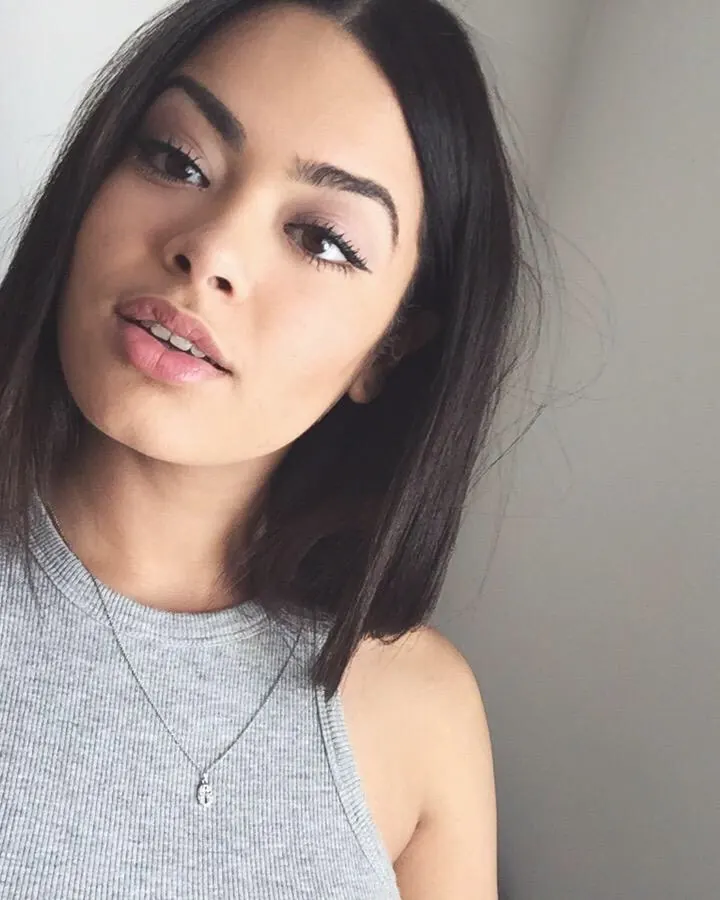 Femme aux cheveux noirs, souriante, vêtue d’un haut gris, posant de profil contre un mur blanc.