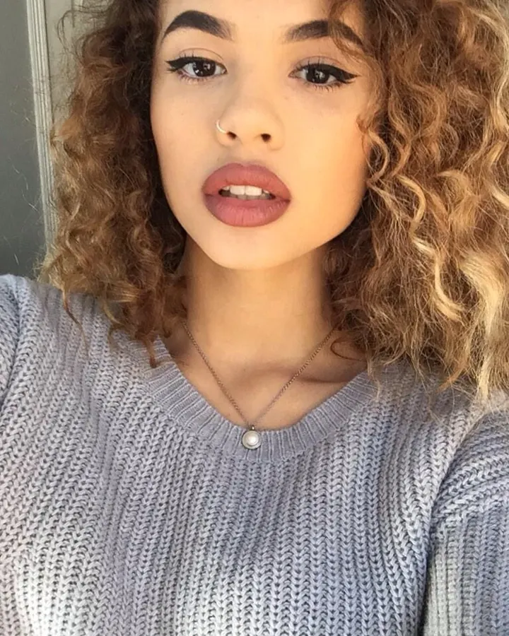 Femme aux cheveux blonds bouclés, souriante, vêtue d’un pull gris clair, posant en plan rapproché.