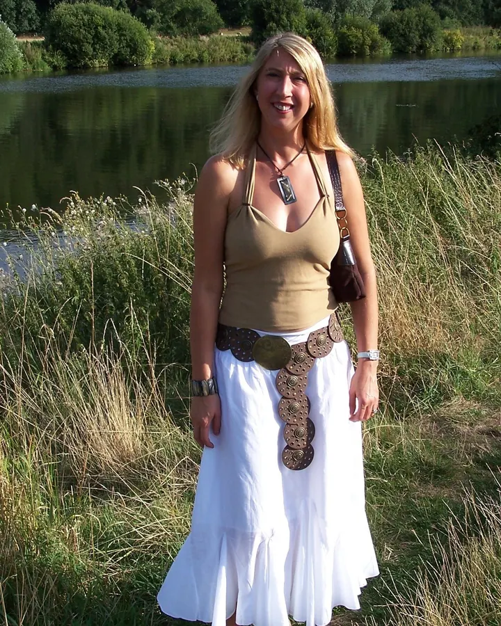 Femme blonde aux cheveux longs, en tenue noire complète avec bottes hautes, appuyée contre un mur de pierre dans une forêt.