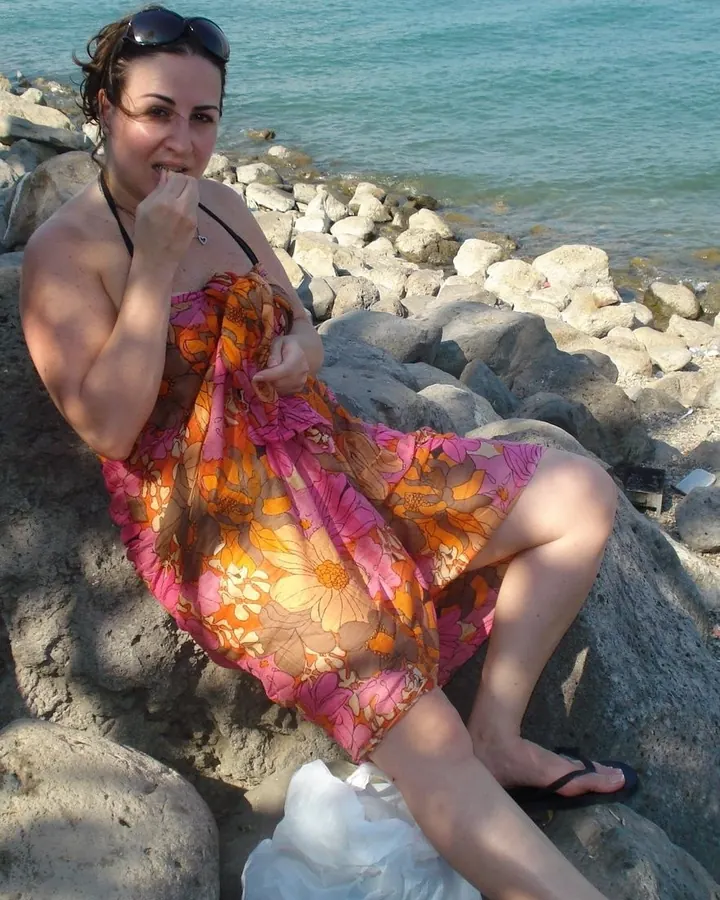 Femme en robe colorée, assise sur des rochers près de la mer, tenant sa robe, lunettes de soleil sur la tête.