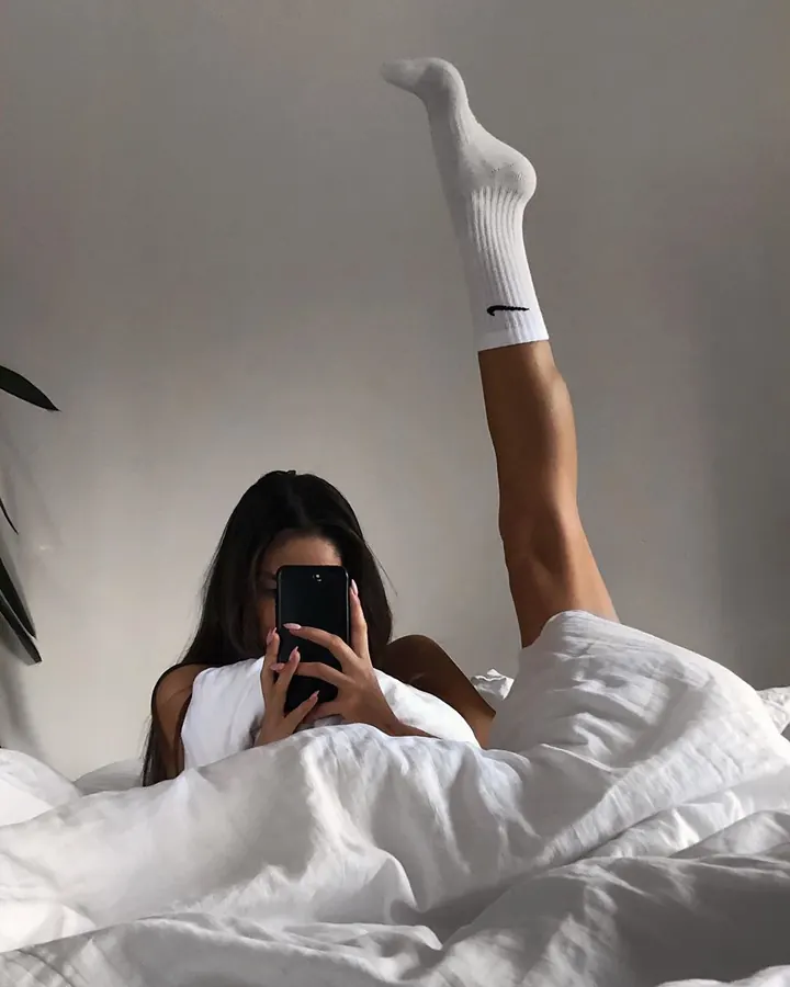 Femme aux cheveux noirs, allongée dans un lit blanc, pied levé en chaussette blanche Nike, prenant un selfie.