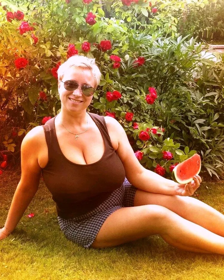 Femme aux cheveux gris courts, portant un haut marron et une jupe à carreaux, assise sur l'herbe devant un buisson de roses rouges, tenant une pastèque.