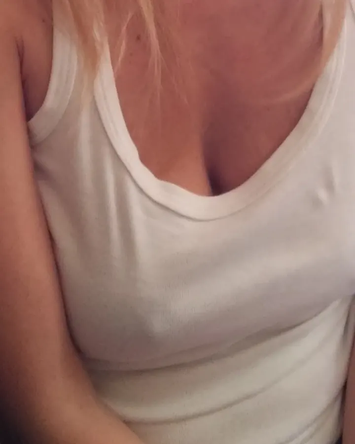 Femme blonde aux cheveux courts, portant un haut blanc et une jupe noire, assise sur un canapé marron en tenant un verre de vin.