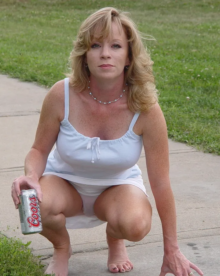 Femme aux cheveux blonds, en tenue blanche, agenouillée sur un trottoir, tenant une canette de Coors, regardant l'appareil.