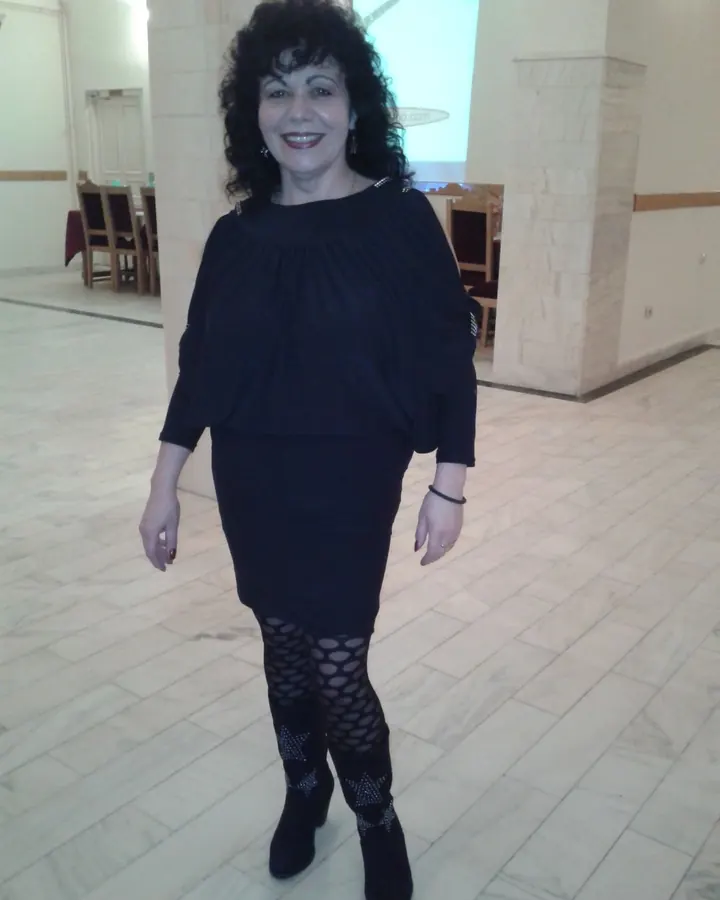 Femme aux cheveux noirs bouclés, en robe noire et bottes, souriante dans une salle avec des tables et des chaises.