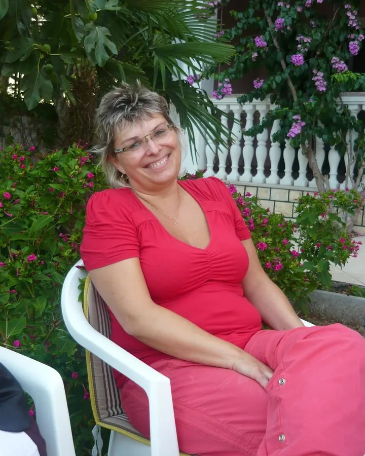 Femme âgée aux cheveux blonds, souriante, robe rouge, assise sur chaise blanche, jardin fleuri en arrière-plan.