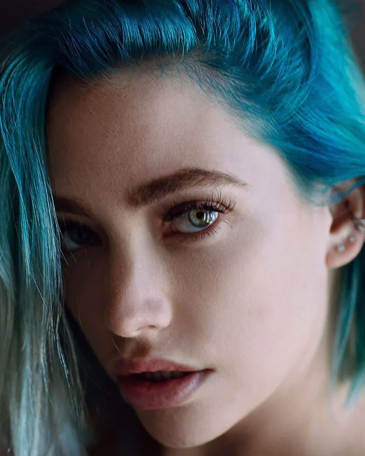 Portrait serré d'une jeune femme aux cheveux bleus, regardant l'appareil, avec un piercing à l'oreille, le visage légèrement ombragé.