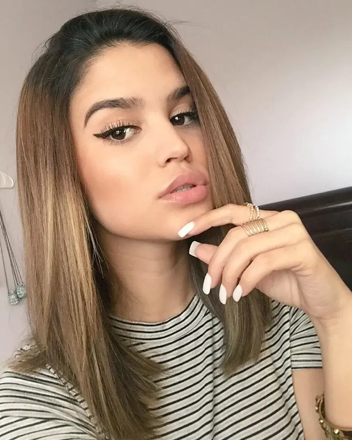Jeune femme aux cheveux châtains en haut noir et pantalon blanc, posant de face, main sur la taille.