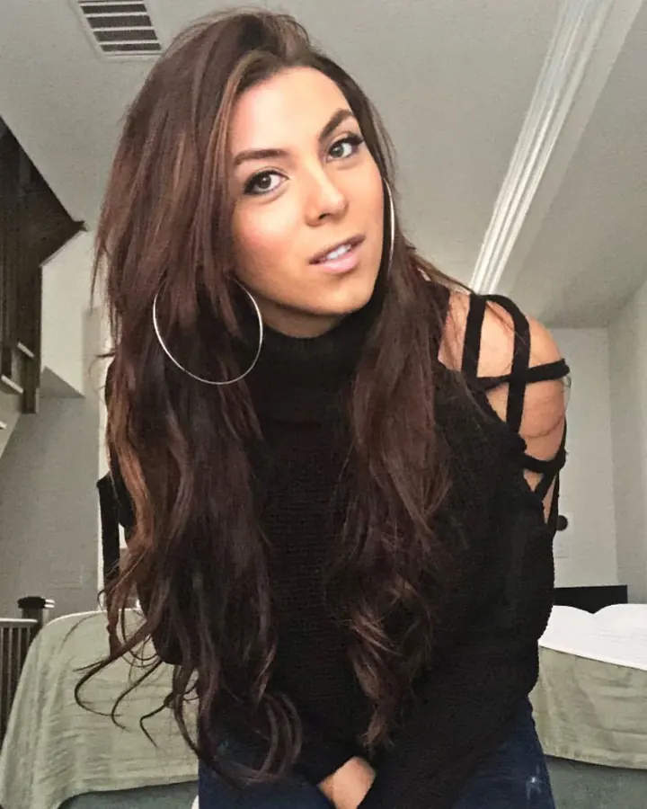 Femme brune en haut noir, posant contre un mur blanc, regardant l'appareil photo