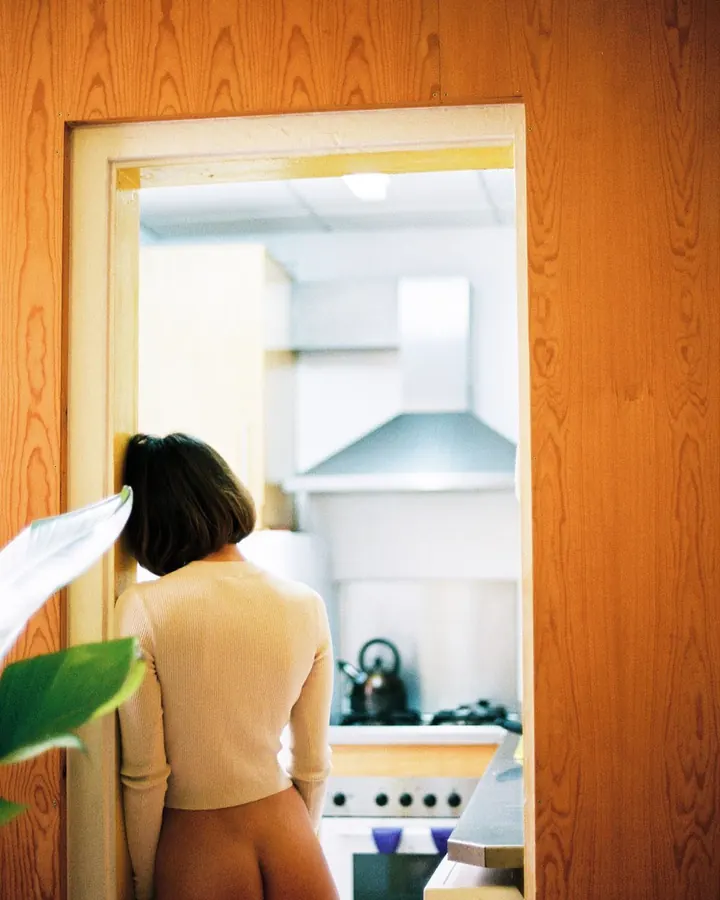 Femme de dos, en haut beige, nue en bas, se penchant vers une cuisine, vue à travers une porte en bois.
