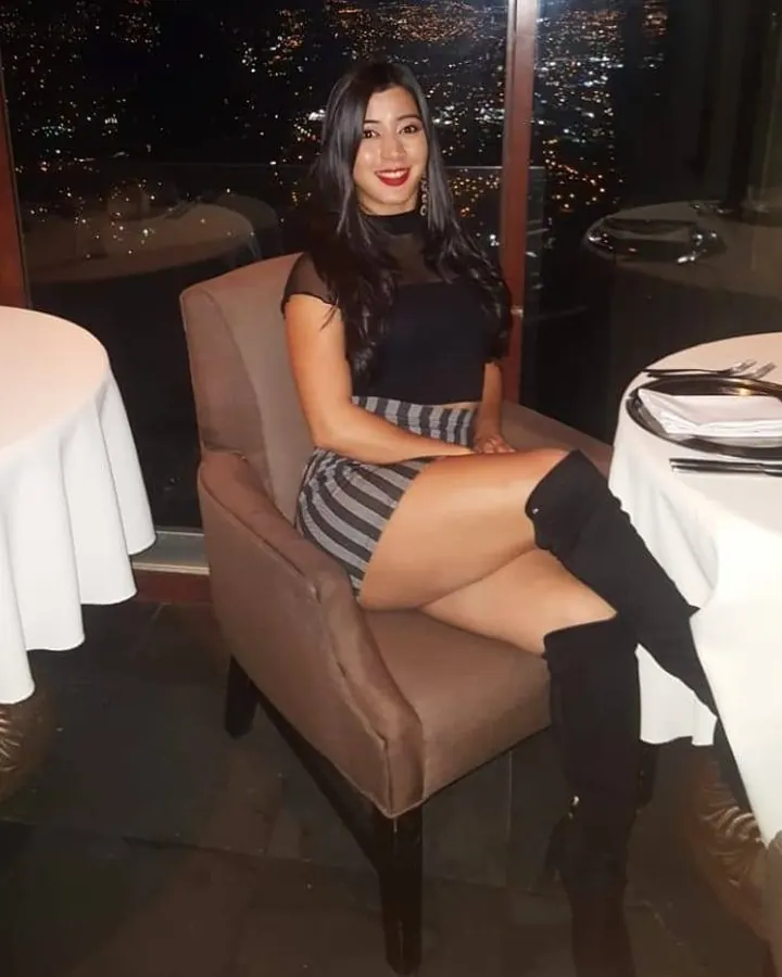 Femme asiatique aux cheveux bruns noirs, souriante, assise dans un fauteuil élégant avec vue sur une ville illuminée la nuit, portant une robe noire et une jupe rayée, avec des bottes hautes.