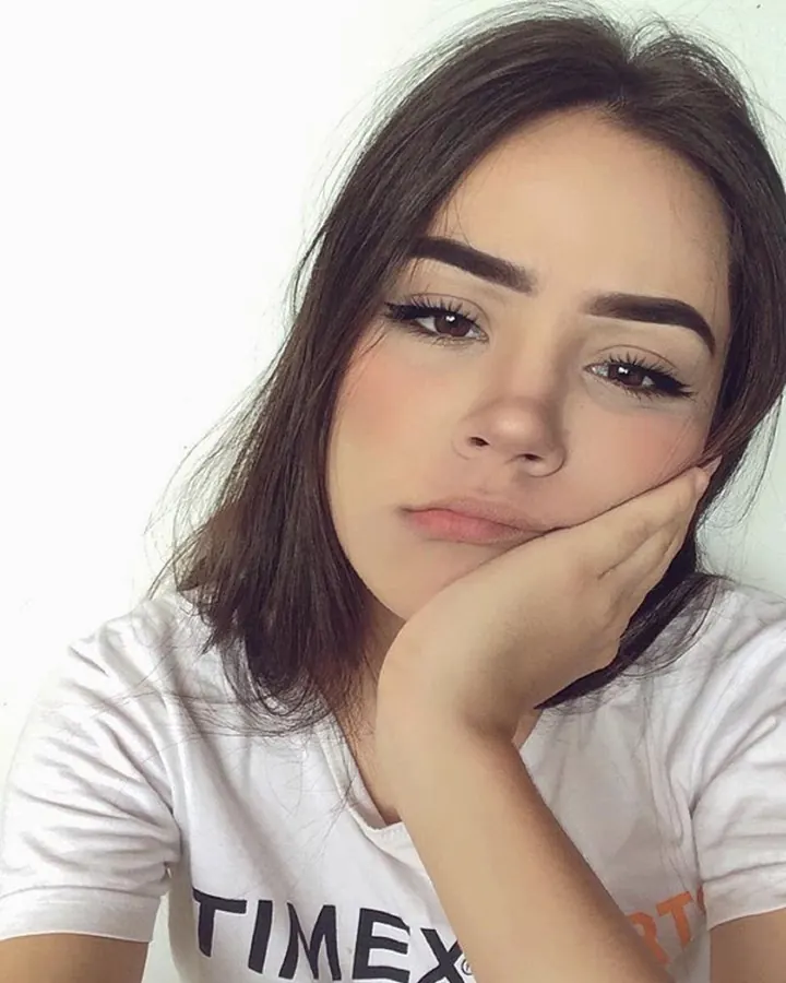 Jeune femme aux cheveux châtains, regard intense, posant près d'un mur blanc, vêtue d'un t-shirt blanc avec l'inscription TIMEX, main sur le menton.