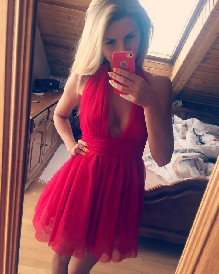 Femme aux cheveux blonds, portant un haut blanc et une jupe plissée à carreaux, tenant un skateboard, debout devant un fond rouge.