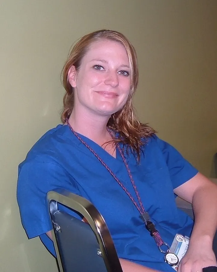 Femme en tenue médicale bleue, assise sur une chaise dans une salle avec un badge autour du cou