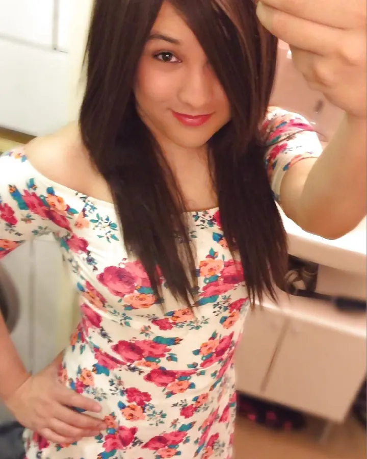 Jeune femme aux cheveux châtains, vêtue d'une robe fleurie blanche, prenant un selfie dans un miroir.