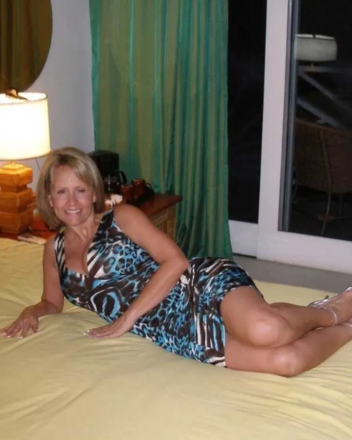 Femme mûre aux cheveux blonds, vêtue d'une robe imprimée bleue et noire, allongée sur un lit jaune dans une chambre éclairée.