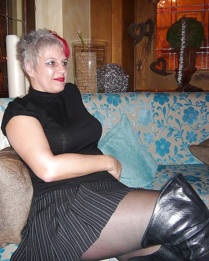 Femme mûre aux cheveux gris, vêtue d'un haut noir, assise sur un canapé à motifs floraux bleus, portant des bottes noires.