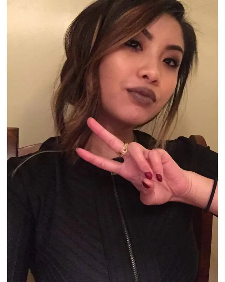 Femme asiatique en tenue sombre, faisant un signe de la main, regard intense, cheveux mi-longs.