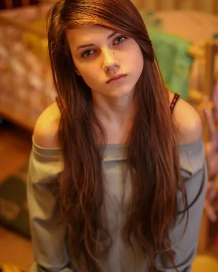 Femme en haut gris, cheveux longs bruns, regardant directement l'appareil, dans une chambre.