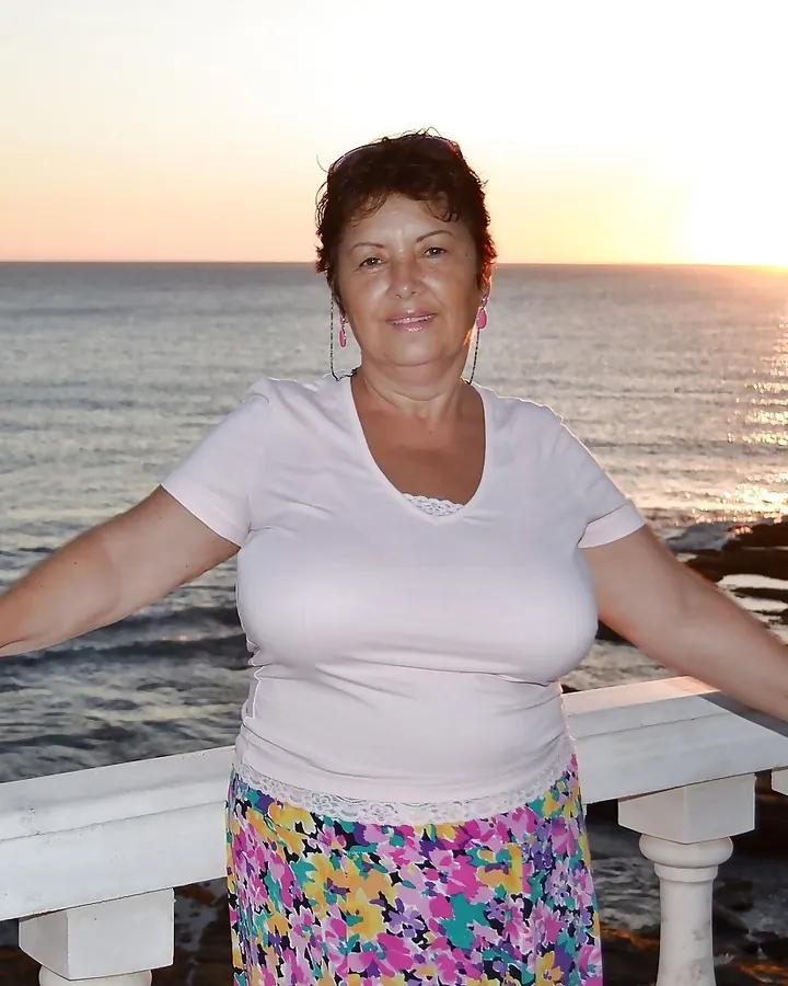 Femme en robe blanche, debout sur un balcon face à la mer au coucher du soleil, bras ouverts.