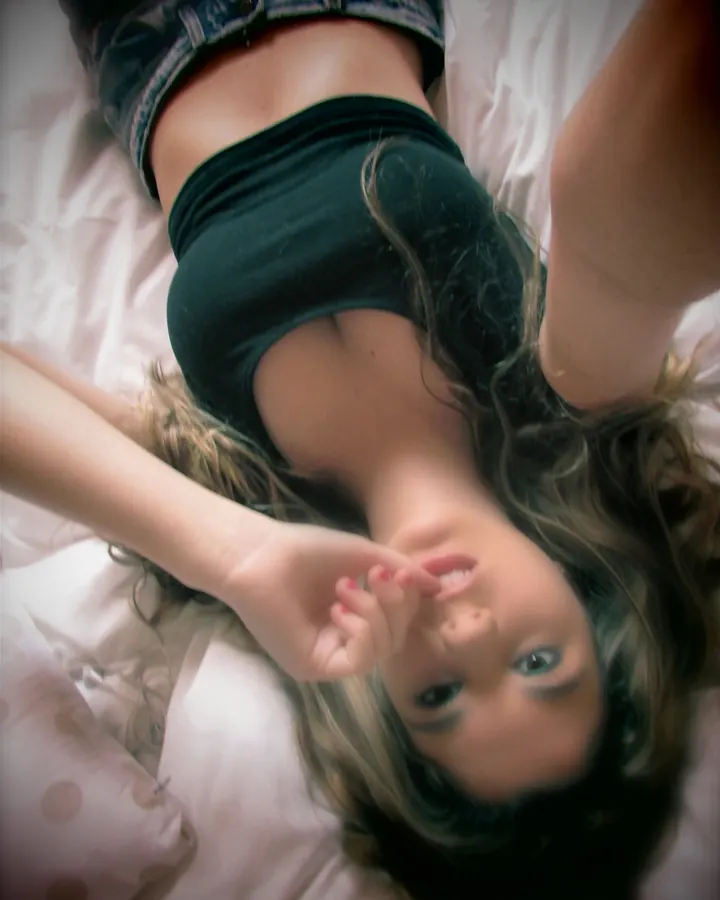 Femme allongée sur un lit, regardant vers le haut, cheveux foncés, vêtue d'un haut noir et de shorts en jean.
