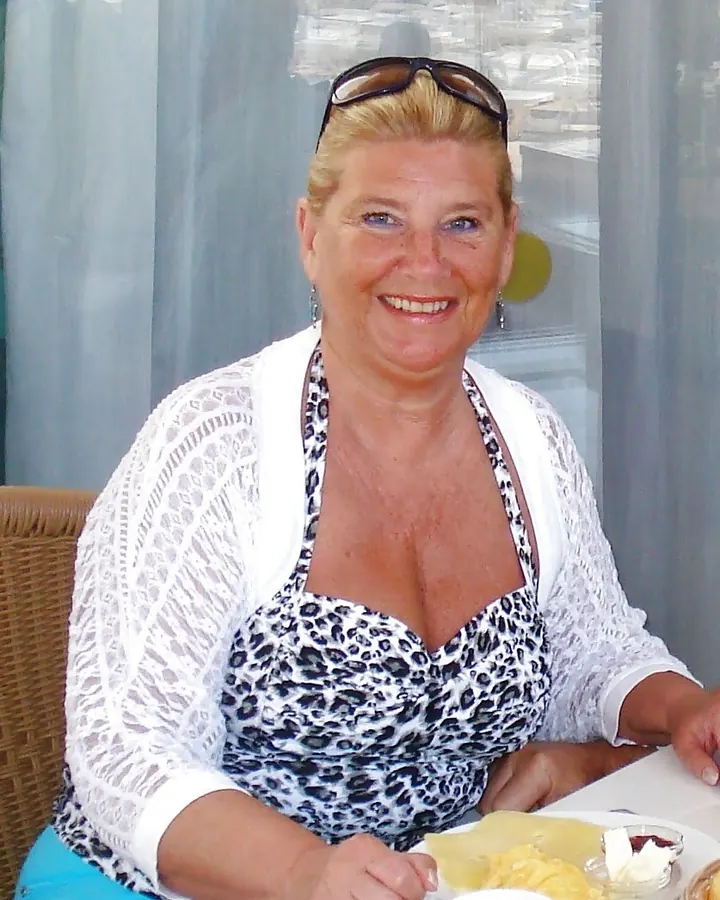 Femme blonde aux yeux bleus, posant dans une cuisine, portant un haut blanc et une culotte violette.