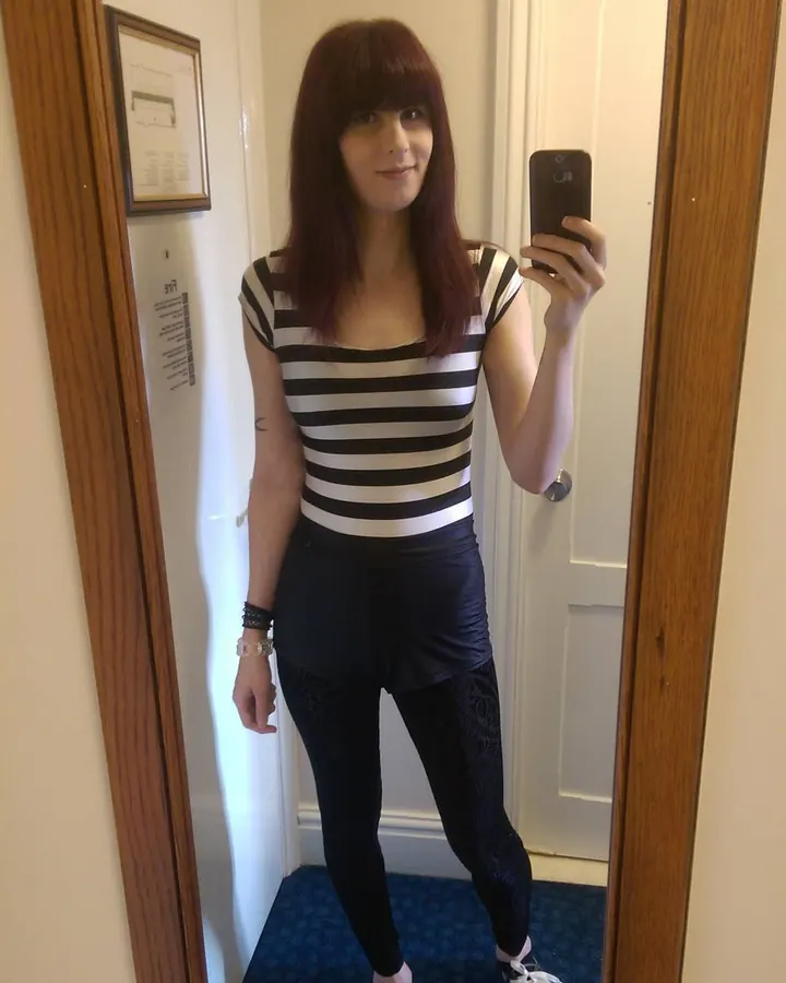 Femme aux cheveux châtains et yeux verts, posant dans une chambre, portant un haut rayé et des leggings noirs.