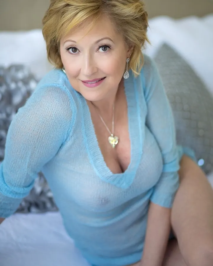Femme aux cheveux blonds, portant un haut bleu clair transparent, assise sur un lit blanc, regardant vers l'appareil photo avec un sourire doux.