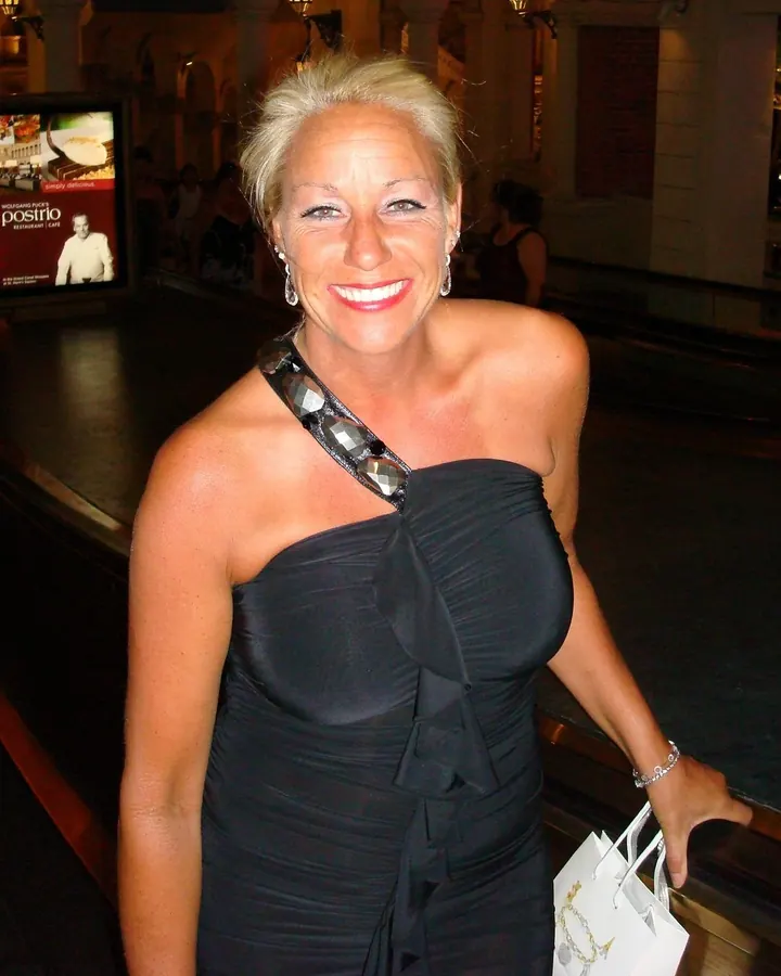 Femme blonde en robe noire asymétrique avec bijoux, souriante dans un hall éclairé.
