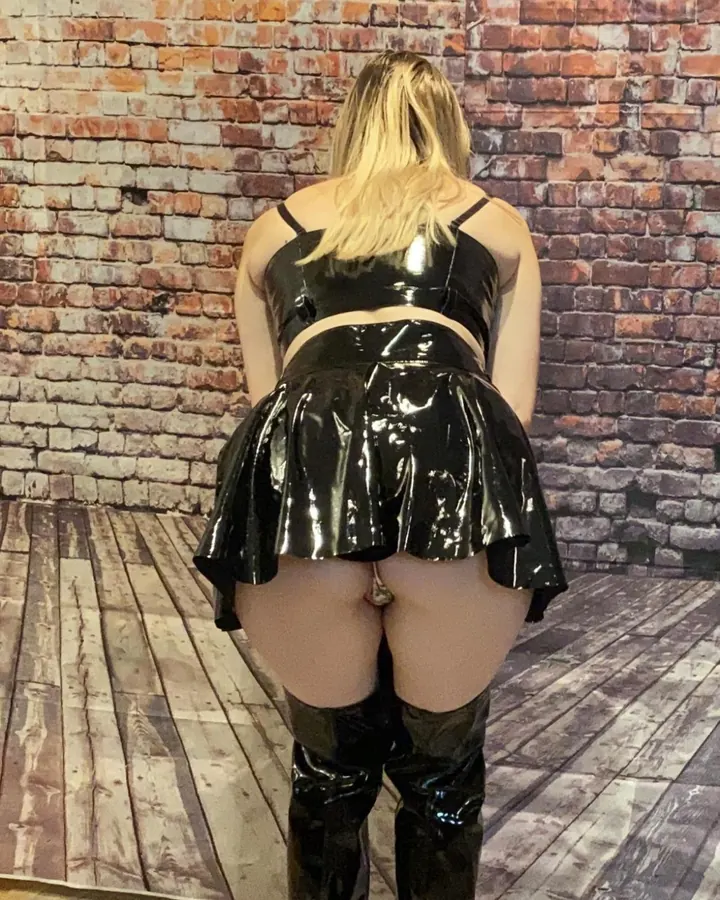 Femme blonde en combinaison noire brillante, posant en squat sur un fond blanc