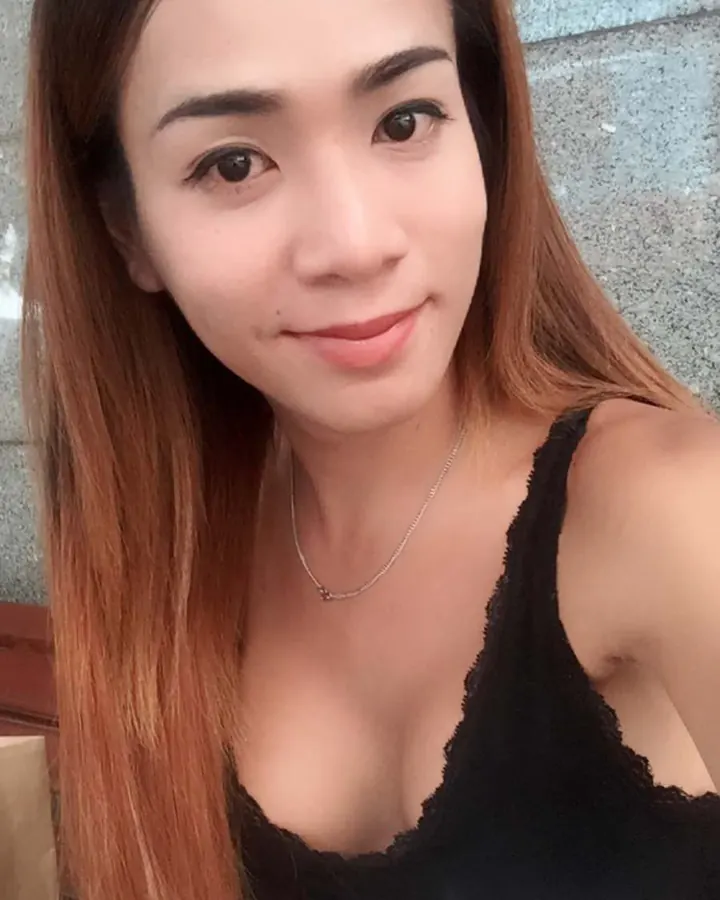 Femme aux cheveux châtains, prenant un selfie, portant un haut noir à dentelle, devant un mur gris.