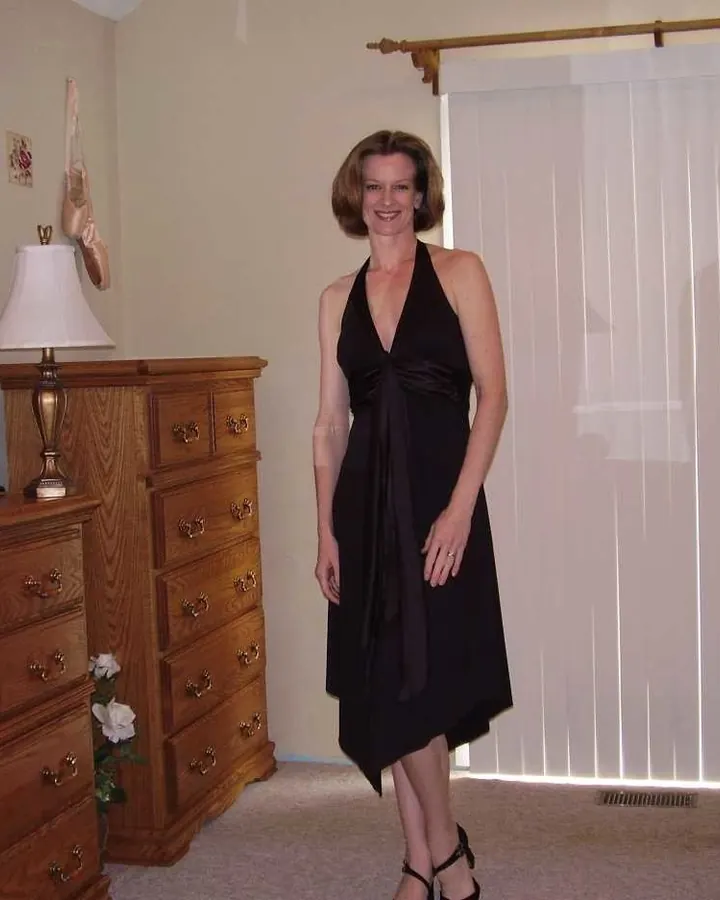 Femme aux cheveux bruns courts, souriante, vêtue d'une robe noire sans manches, debout dans une chambre avec un meuble en bois.