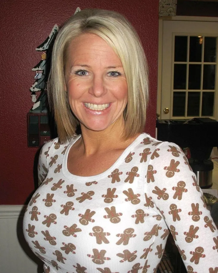 Femme aux cheveux blonds, souriante, portant un haut blanc, un collier, des collants noirs, des chaussures rouges, assise sur une chaise dans une chambre.