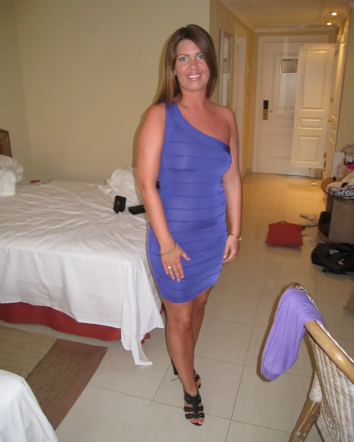 Femme souriante en robe bleue asymétrique, dans une chambre avec lit blanc et sol carrelé.