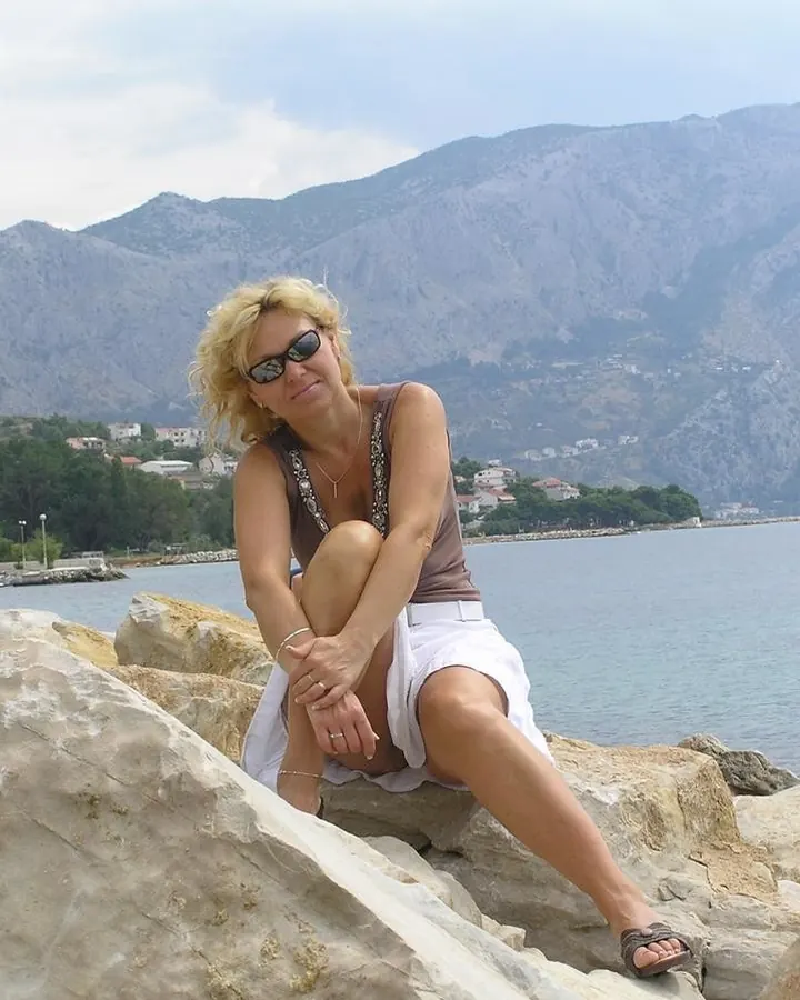 Femme blonde assise sur des rochers près de la mer, portant un haut marron et une jupe blanche, avec des lunettes de soleil.