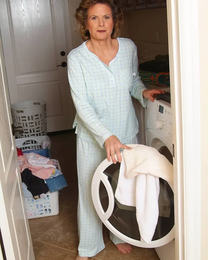 Femme en pyjama à carreaux clairs, pieds nus, tenant une serviette devant une machine à laver, dans une buanderie.