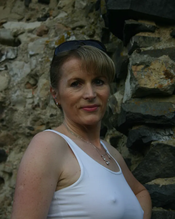 Femme aux cheveux blonds, portant un débardeur blanc, debout devant un mur de pierres, regardant l'appareil photo.