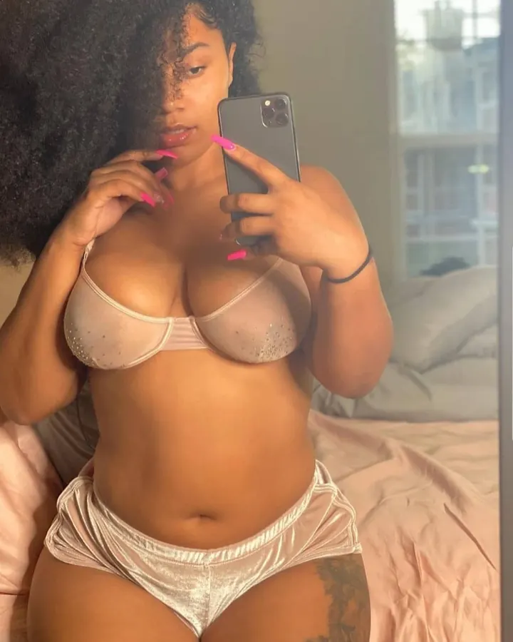 Femme aux cheveux crépus, portant un ensemble de lingerie rose pâle, prenant un selfie devant un miroir, avec un tatouage sur la cuisse.