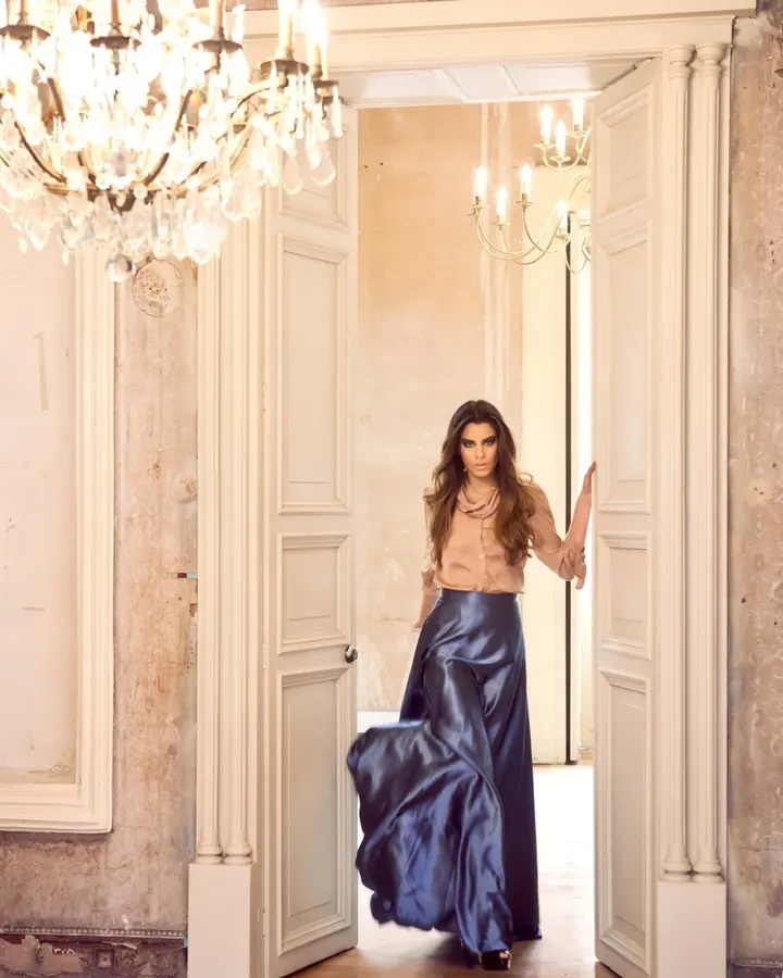 Femme brune en chemise beige et jupe bleue satinée, marchant dans un hall lumineux sous un lustre.