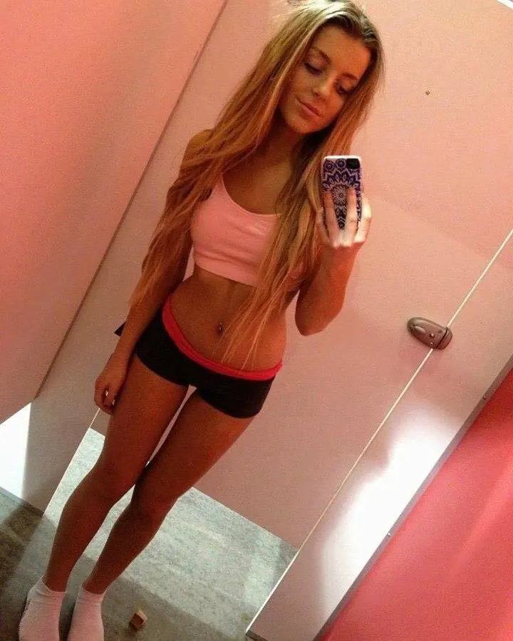 Femme aux cheveux blonds portant un haut rose et un short noir, prenant un selfie dans un miroir, regardant l'objectif avec un sourire.