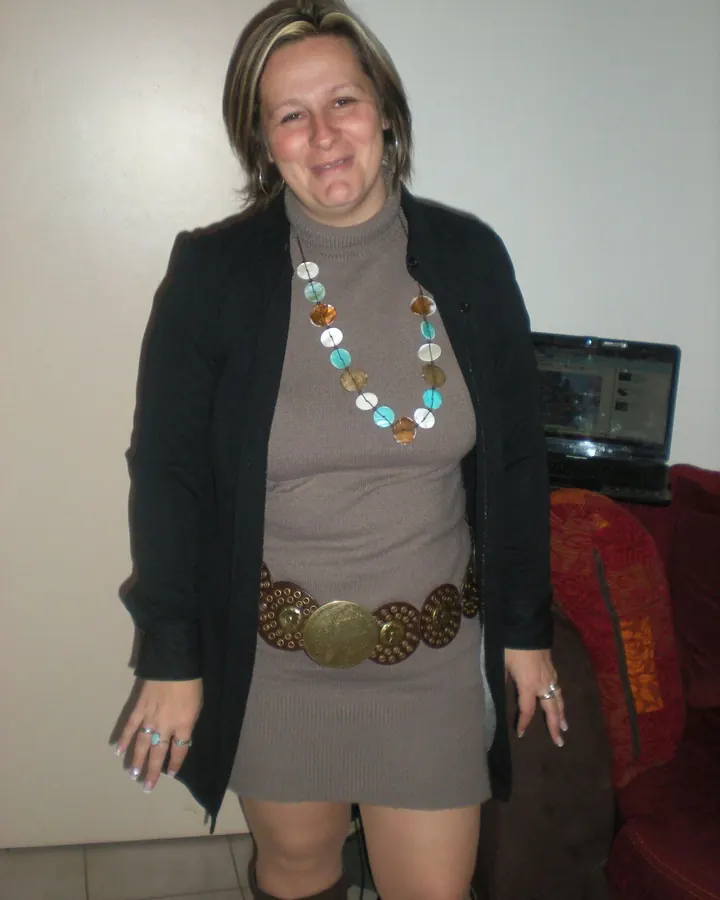 Femme souriante, portant une robe grise, une veste noire et un collier de pièces colorées, debout dans un salon avec un ordinateur portable en arrière-plan.
