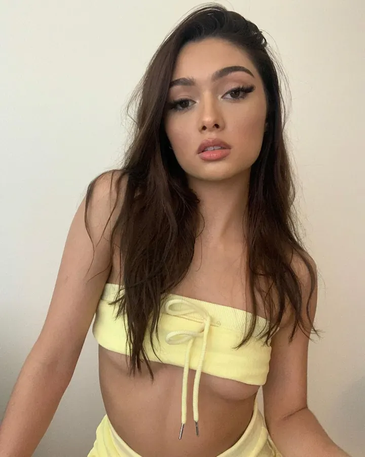 Femme aux cheveux bruns, portant un haut jaune fluo avec un lien, regardant directement l'appareil photo avec un maquillage soigné.