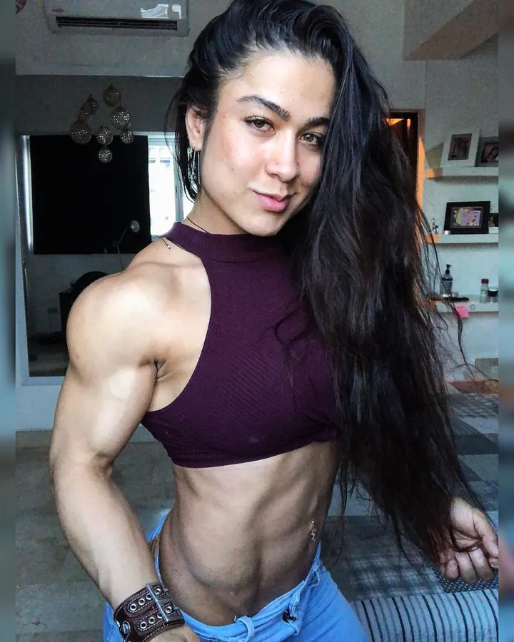 Femme aux cheveux bruns, en haut bordeaux, muscles visibles, jeans, posant dans un intérieur moderne