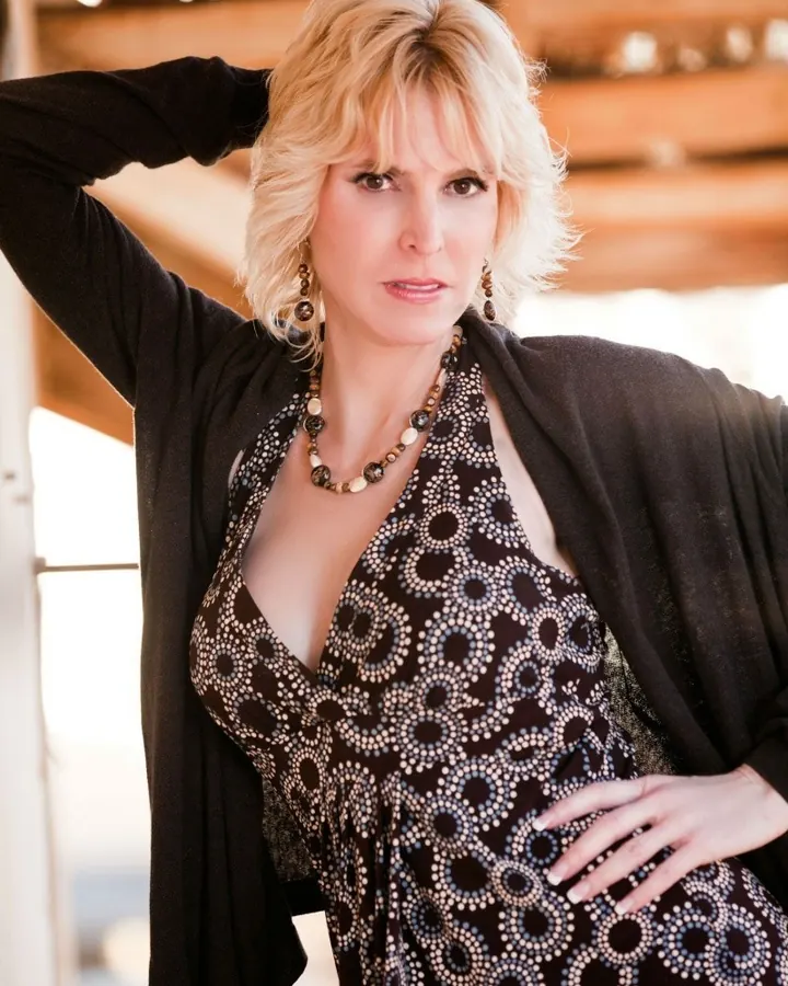 Femme aux cheveux blonds, portant une robe noire à motifs circulaires, avec un cardigan noir, pose en extérieur sous une structure en bois.