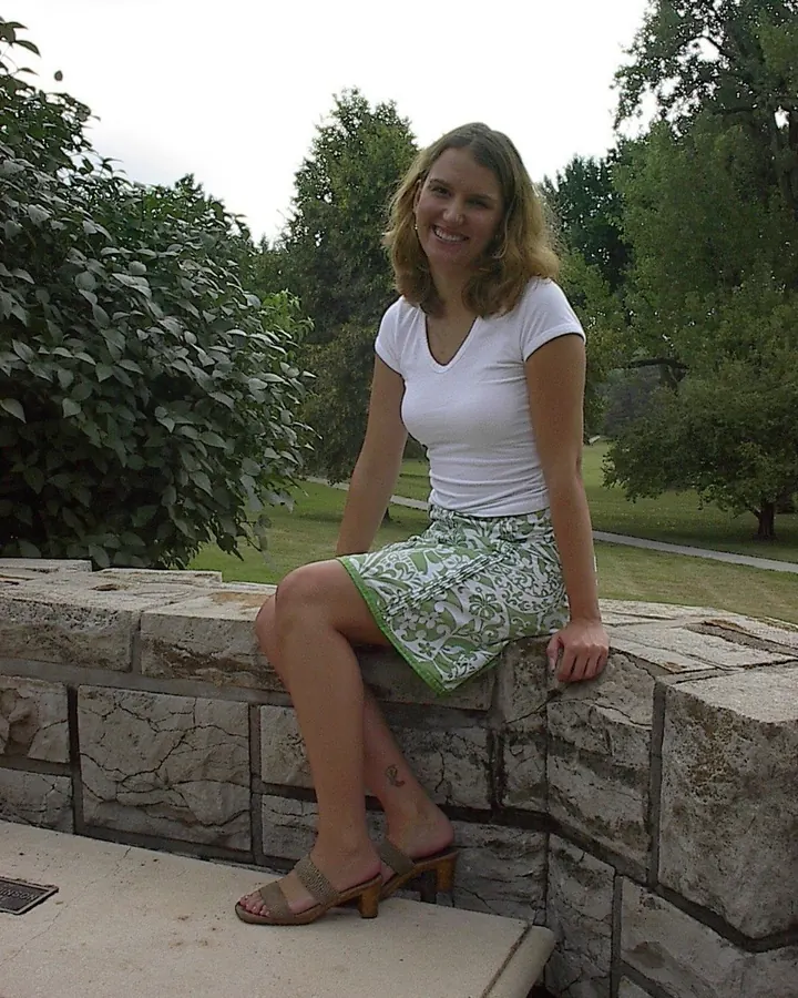 Femme aux cheveux châtains, portant une robe noire sans manches, debout contre un arbre dans un jardin, avec des bottes en cuir noir et un tatouage floral sur la jambe.