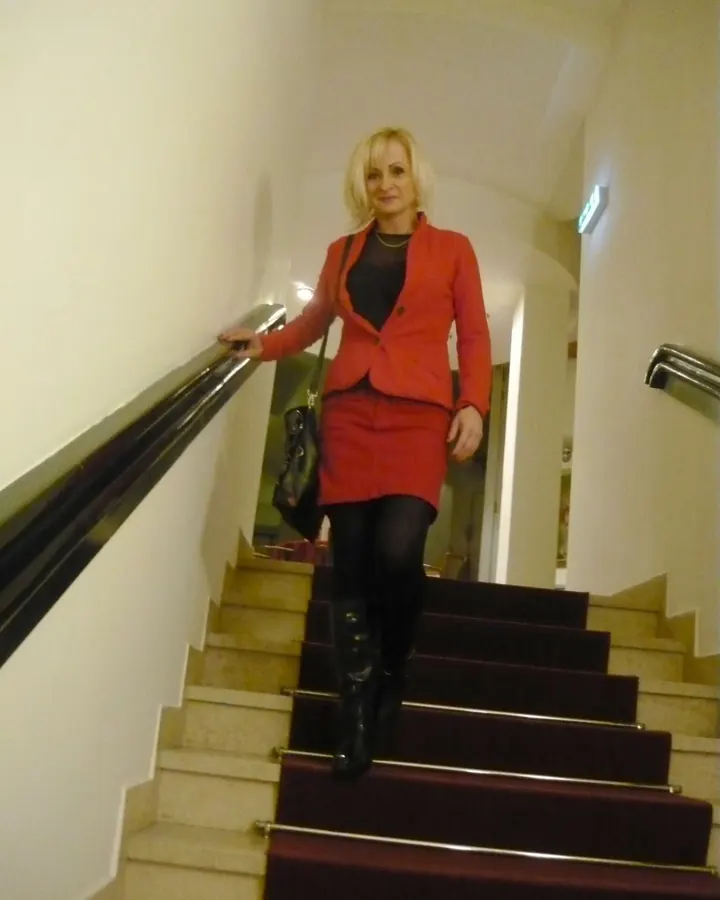 Femme blonde aux yeux marrons, vêtue d'une veste rouge et d'une jupe assortie, descendant un escalier avec une main sur la rampe.