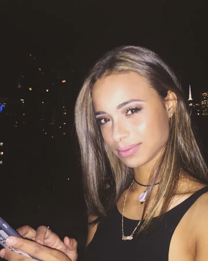 Femme aux cheveux bruns, en haut noir et collier, posant de nuit avec une ville illuminée en arrière-plan, tenant un téléphone.