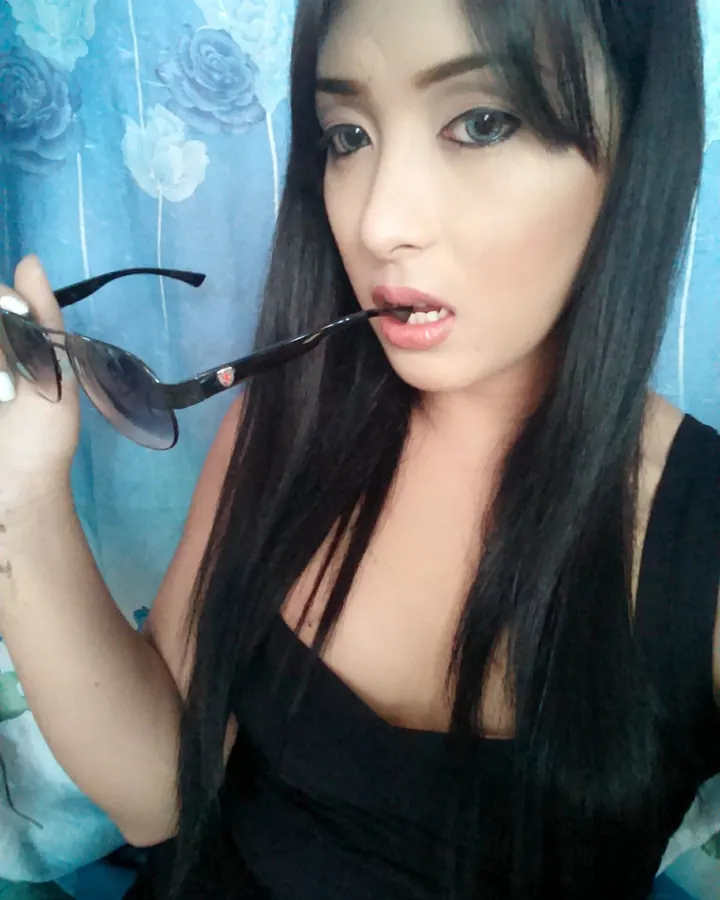 Femme asiatique aux cheveux noirs, en haut noir, tenant des lunettes de soleil près de sa bouche, devant un fond bleu floral