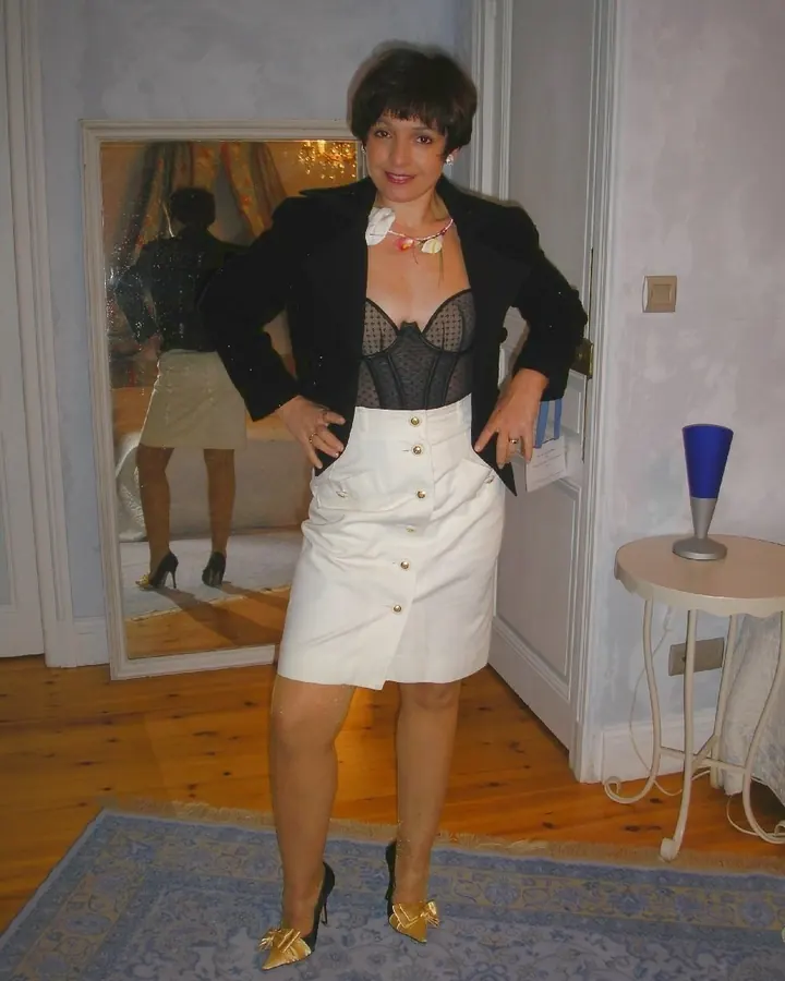 Femme aux cheveux bruns courts, vêtue d'une veste noire, d'un corset noir et d'une jupe blanche boutonnée, posant dans une pièce élégante avec un miroir en arrière-plan.