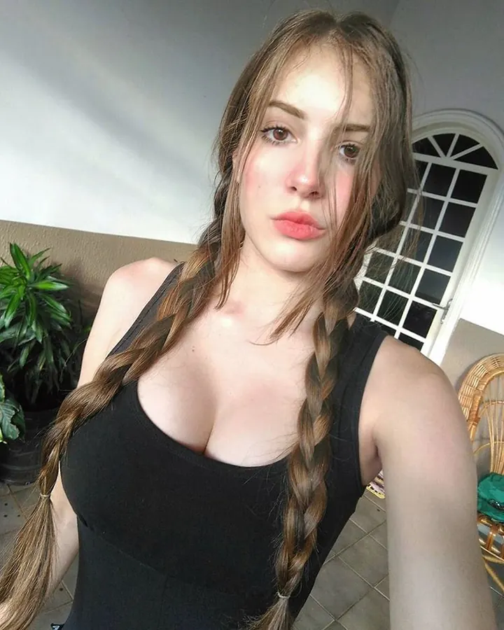 Jeune femme aux cheveux bruns en tresses, vêtue d'un haut noir, regardant l'appareil photo dans un intérieur lumineux.